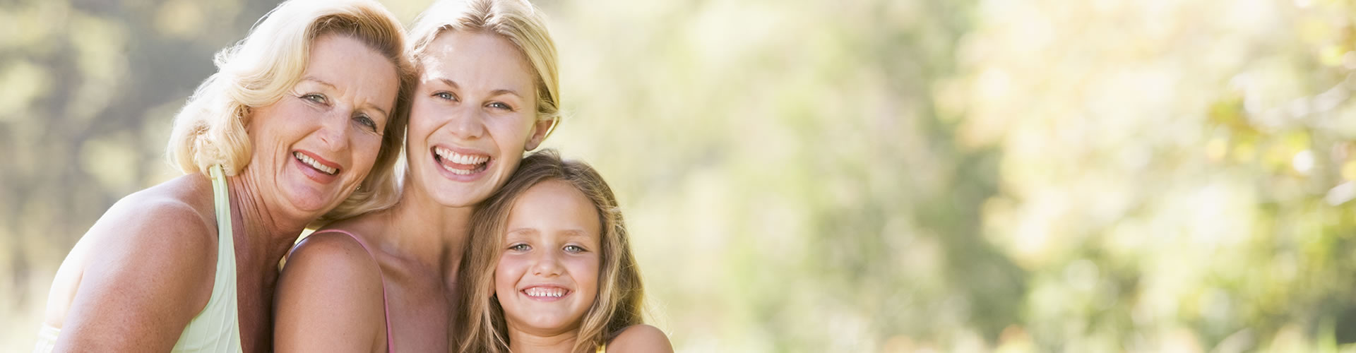Three generations of women—an older woman, a younger woman, and a young girl—smiling outdoors with trees and sunlight in the background as they commemorate the young girl’s first visit to this special place.