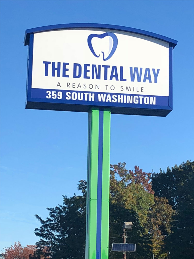A sign reads THE DENTAL WAY, A REASON TO SMILE, 359 SOUTH WASHINGTON with a tooth logo above. The sign is mounted on a green pole against a blue sky and trees, welcoming visitors and letting new patient forms streamline your first visit.
