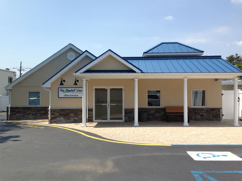A dentist office with a blue roof, beige walls, a front glass door, a bench, and a sign reading The Dental Group Office Entrance. A handicap parking spot is visible in the foreground, and new patient forms are available for easy access as you enter.