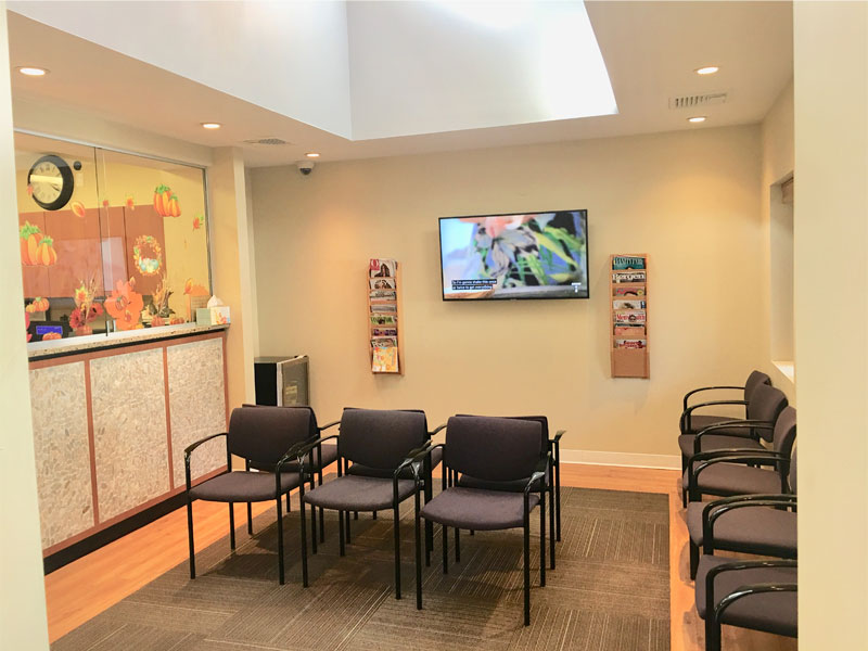 A waiting room with several empty chairs, a TV on the wall, brochures and new patient forms in holders, and a reception area decorated with autumn-themed items. Bright lighting and clean, neutral decor.