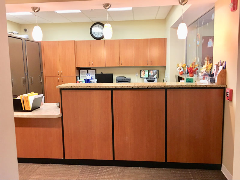 Reception desk with wooden cabinets, countertop with folders, new patient forms, and office supplies, pendant lights above, and fall decorations on the right side. A clock is on the wall above the cabinets in the background.