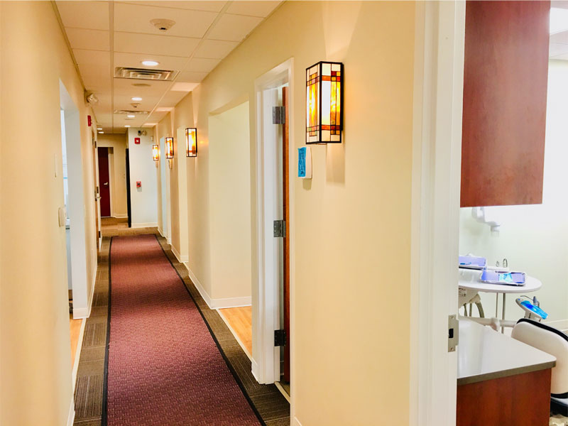 A brightly lit hallway with cream walls and wall lights, leading to several doors. One open door reveals a dental chair and equipment in a treatment room, where new patient forms are ready for your first visit.