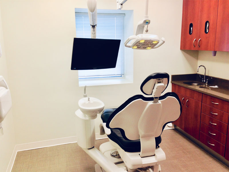 A modern dental exam room with a dentist chair, overhead light, monitor, sink, and wooden cabinets. A window with blinds lets in natural light. The setup appears clean and organized, with new patient forms conveniently placed for your visit.