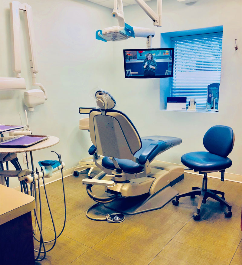 A modern dental office with a dental chair, overhead light, dental tools, a blue stool, and a TV screen displaying a video game. A window with blinds lets in natural light, while new patient forms are neatly arranged at the front desk for easy access.