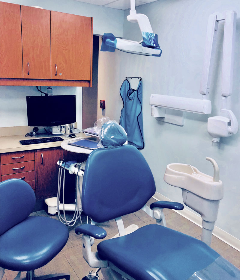 A modern dental exam room with a blue dental chair, sink, computer, cabinets, X-ray machine, and a protective lead apron hanging on the wall is ready to welcome visitors—with new patient forms conveniently available for a streamlined first appointment.